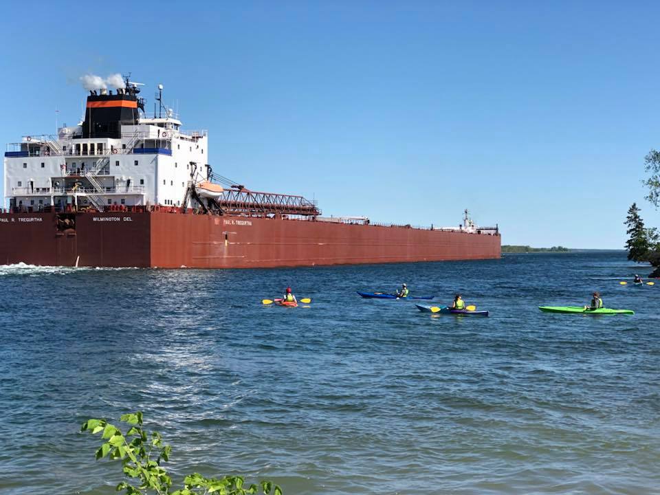 Five kayakers paddling next to a large brown and white freighter in the St. Marys River.