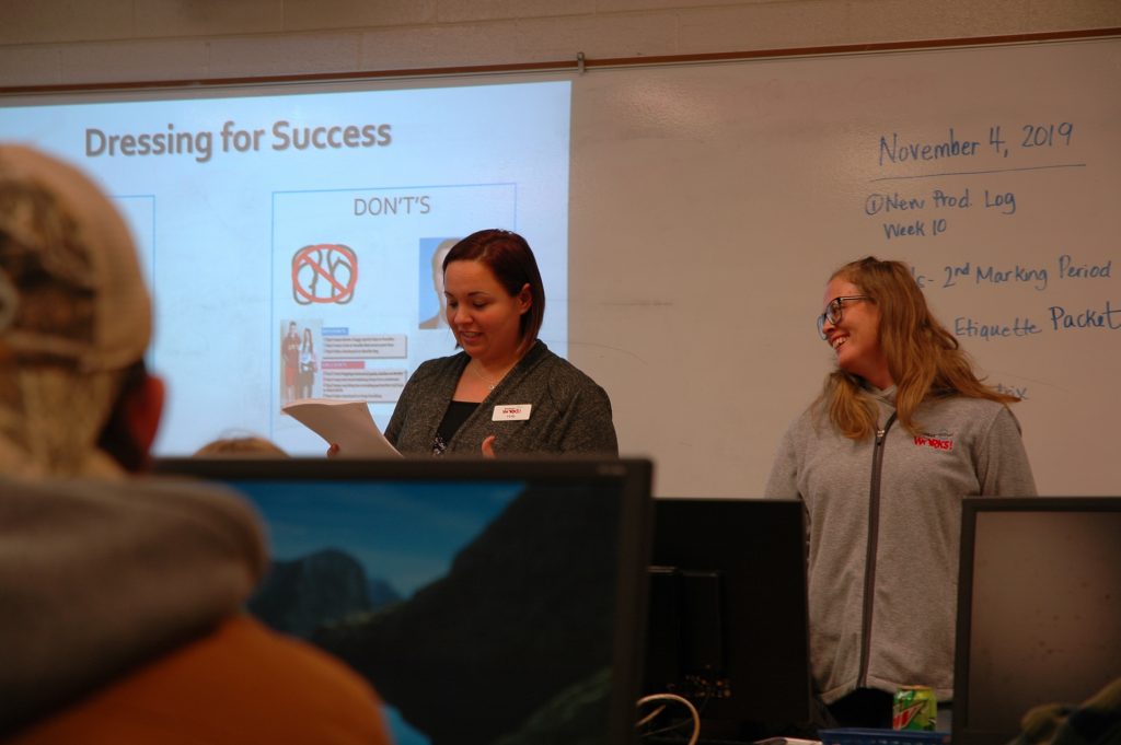 Two Michigan Works! staff members stand in front of the class. One is looking at a piece of paper and the other is laughing. A man with a hat can be see in the foreground.