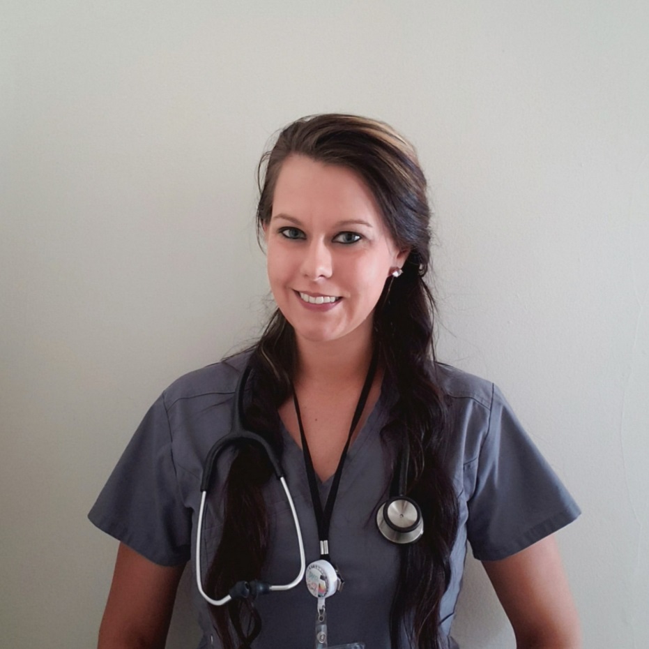 Nichole stands in front of a blank wall wearing gray scrubs, a stethoscope, and a badge.