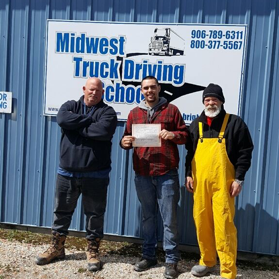 David stands in front of a Midwest Truck Driving School sign with a certificate in his hand, standing between two other men.