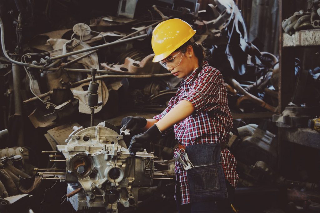 A woman wearing safety equipment works on a piece of machinery in a factory setting.