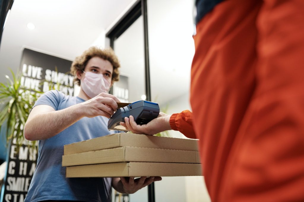 A man in a mask receives pizza from a delivery person while he wears a mask.