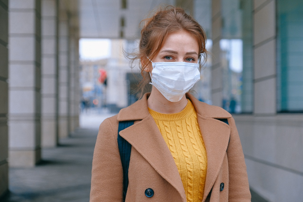 A woman outside in a brown jacket wears a mask.