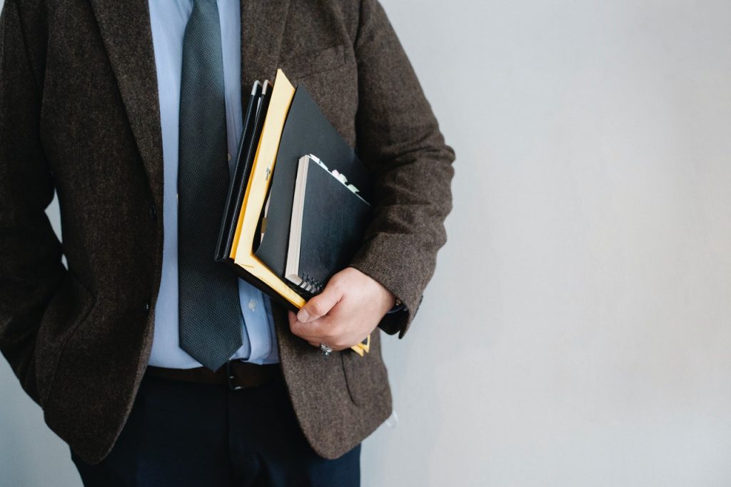 A man in a blazer and tie holds folders and a notebook.