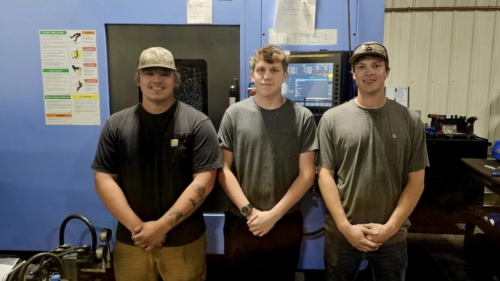 Three students or employees stands in front of a CNC Machine and look into the camera